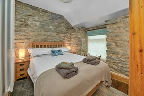 A bedroom with a stone wall, a wooden bed frame, a nightstand, a lamp, and a window with a view. - Charming Snowdonia Slate Cottage (Blaenau Ffestiniog)