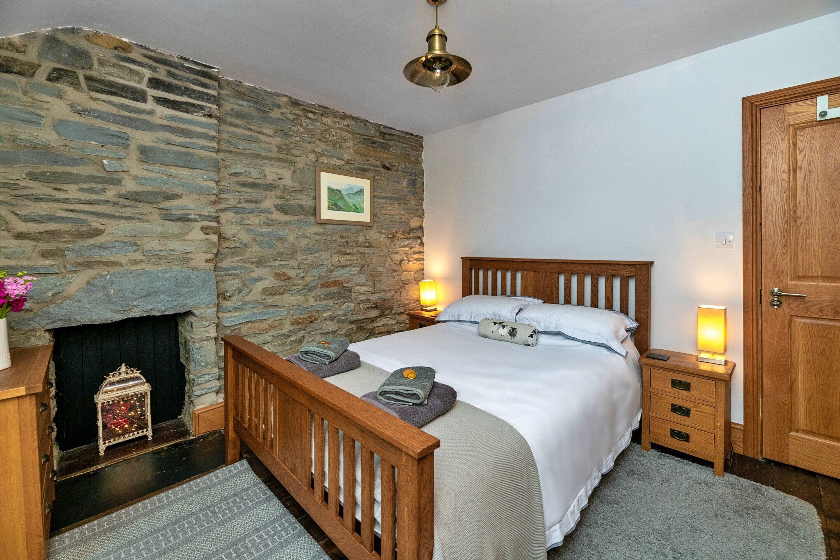 A bedroom with a stone wall, a bed with wooden headboard, a nightstand, a bedside lamp, and a door.