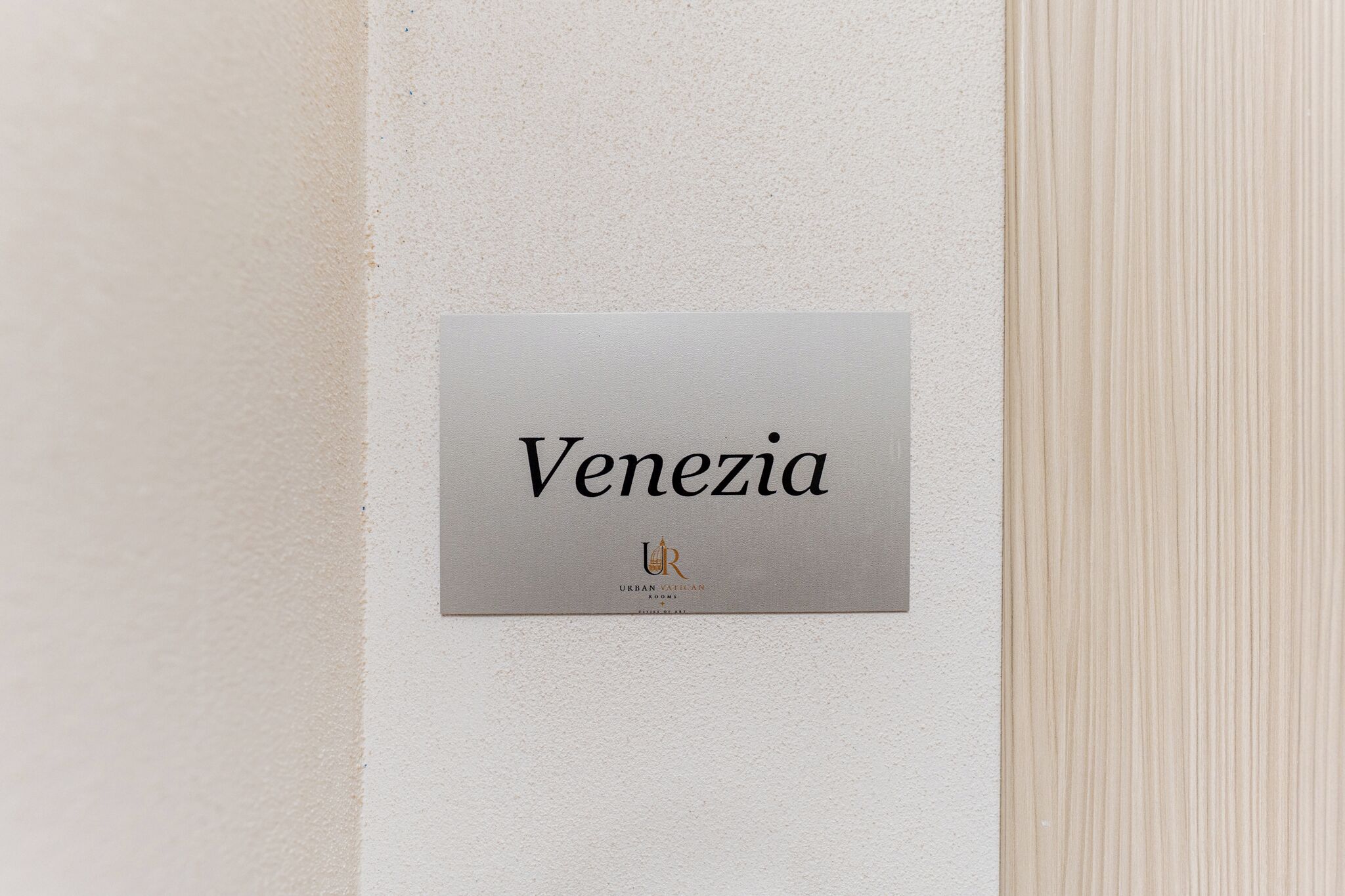 Un letrero con el nombre 'Venezia' y el logotipo 'URBAN VATICAN ROOMS' sobre una pared texturizada.