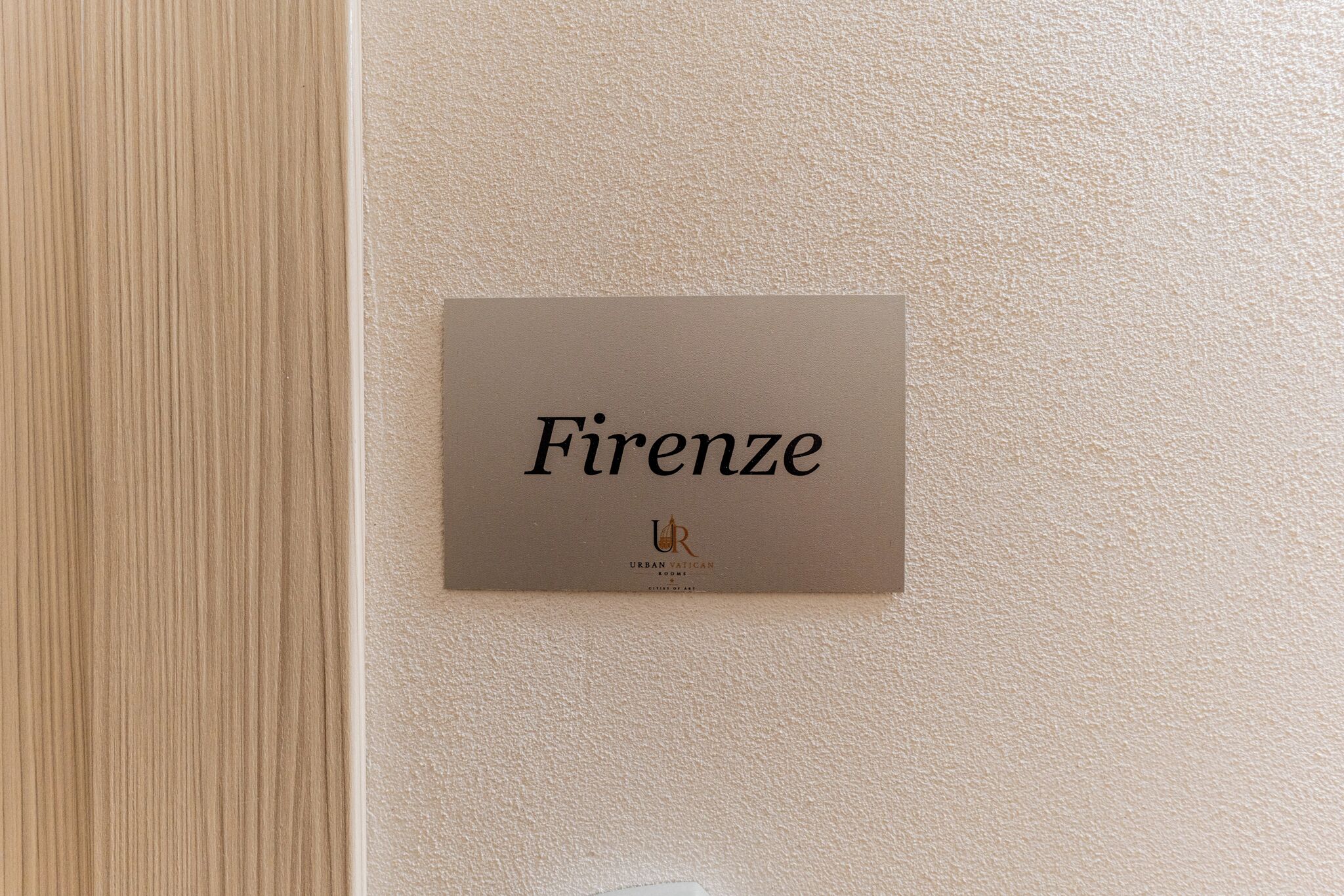 Ein Schild mit dem Wort 'Firenze' und dem Logo von 'Urban Vatican Rooms' an einer strukturierten Wand.
