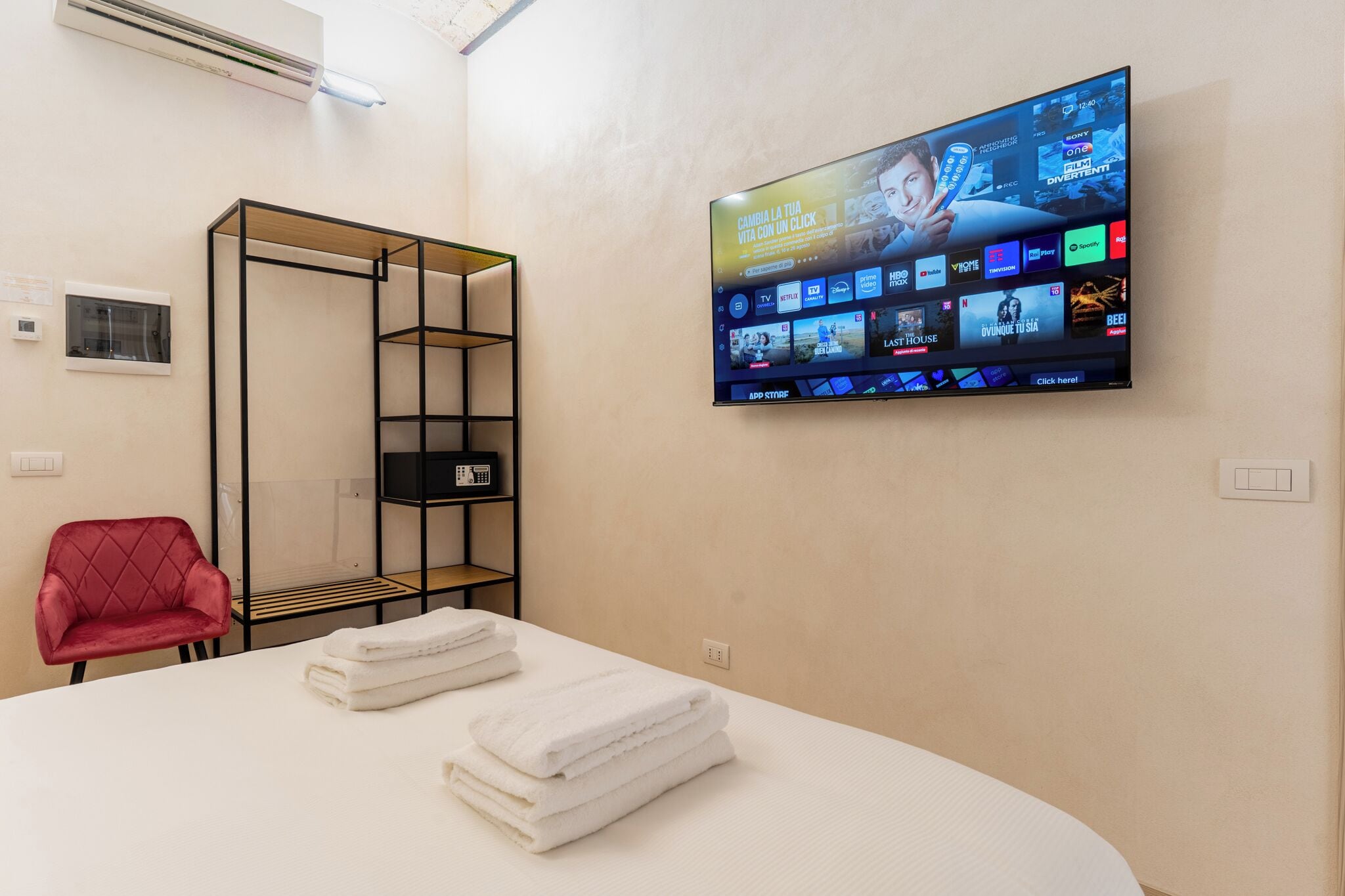 Une chambre d’hôtel équipée d’un téléviseur à écran plat diffusant divers services de streaming, d’un fauteuil rouge, d’une table blanche avec des serviettes et d’une étagère noire.