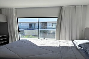 A hotel room with a large bed, a desk, and a view of a building through a glass door. - Studio privativo em BG a 2km da praia (Cajueiro da Praia)