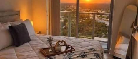 A hotel room with a large bed, a view of the sunset, and a cityscape.
