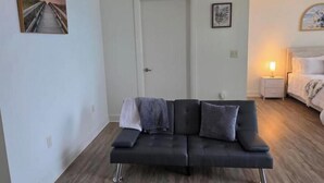 A room with a black sofa, a door, a bedside table with a lamp, and a bed with a white headboard. - Dream Stay (Fort Myers)