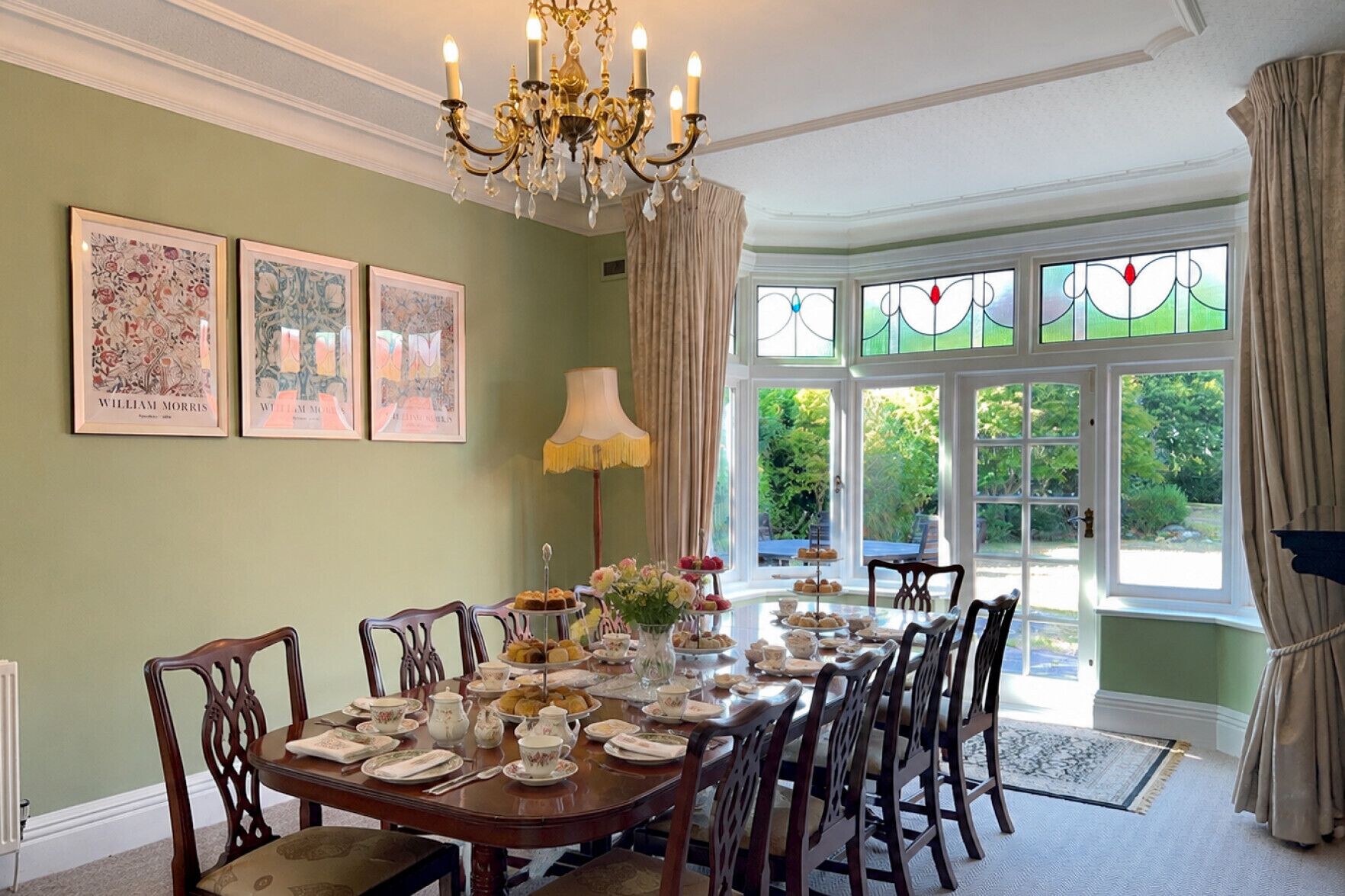 A dining room with a round table set for a meal, surrounded by chairs, with a chandelier above and a window with curtains to the right.