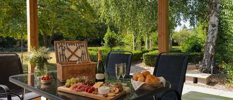 An outdoor dining area with a glass table, wicker picnic basket, and a variety of food and drinks.