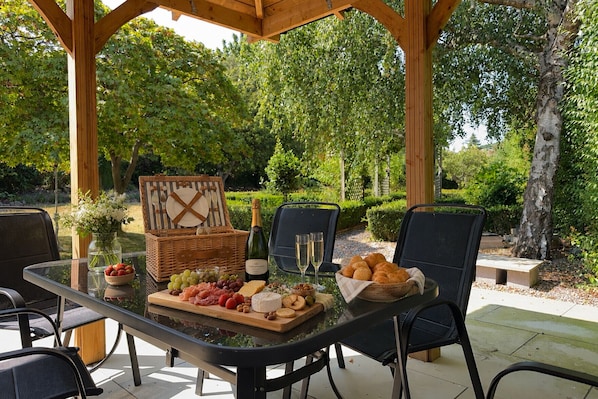 Un espace de restauration en plein air avec une table en verre, un panier de pique-nique en osier et une variété de plats et de boissons.