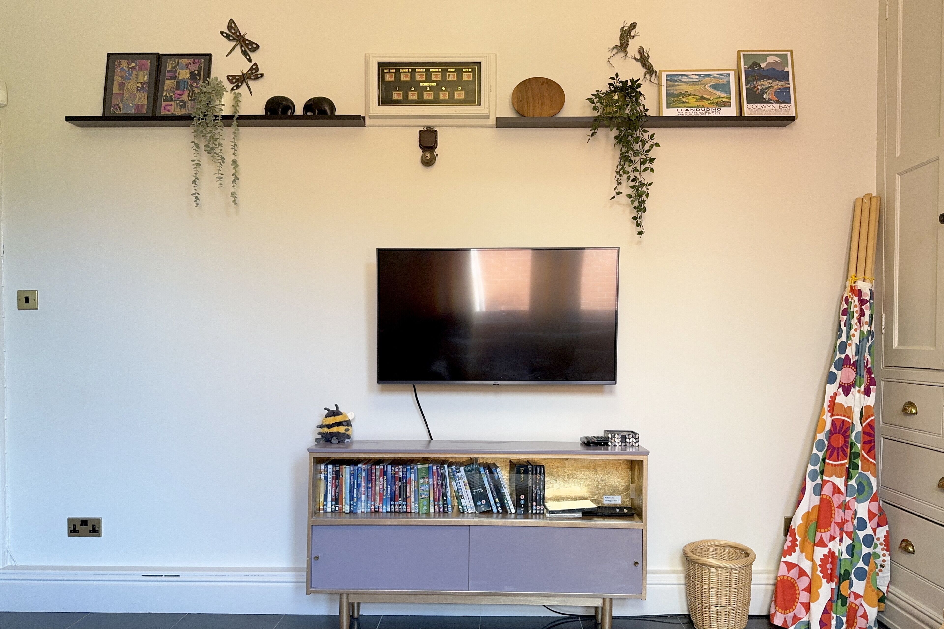 A living room with a flat-screen TV mounted on the wall, a bookshelf with books and decorative items, and a door with a colorful curtain.