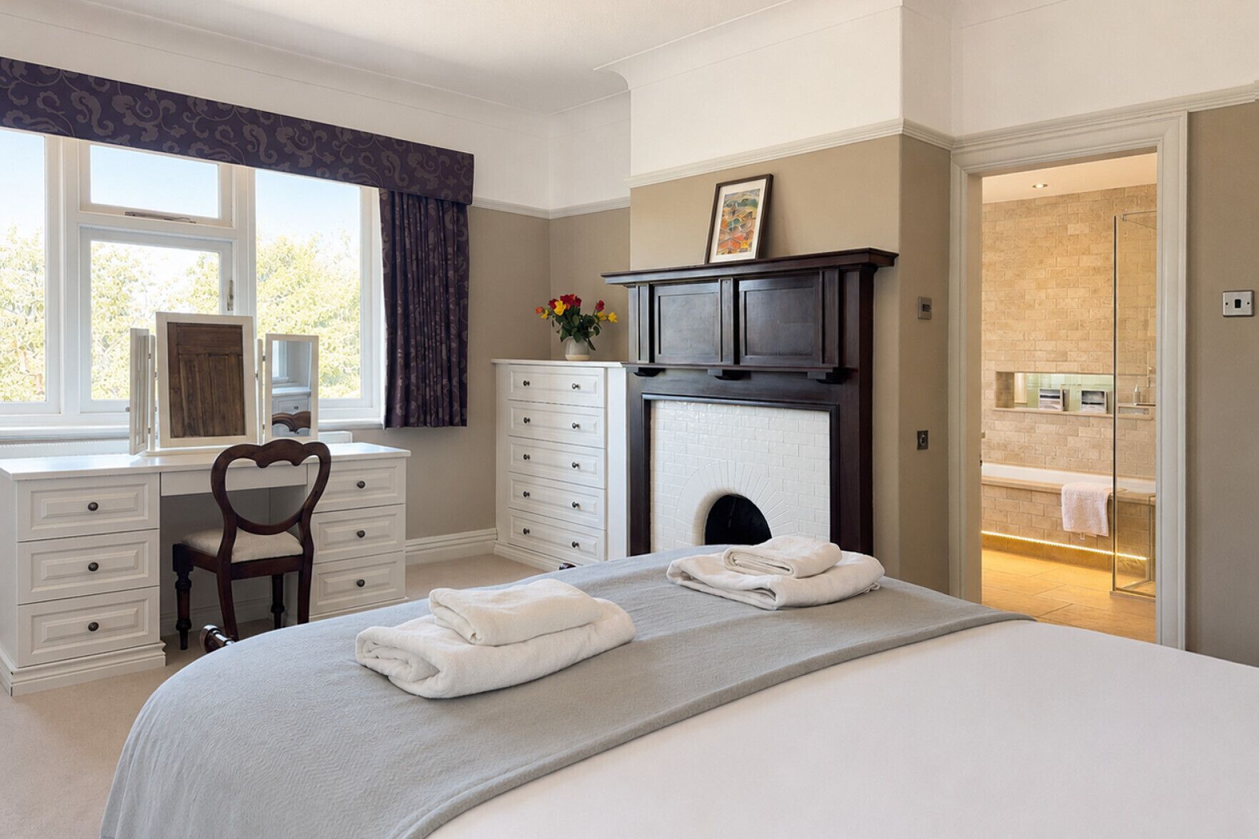 Quarto com cama, cômoda, cadeira, lareira e banheiro visível por uma porta aberta.