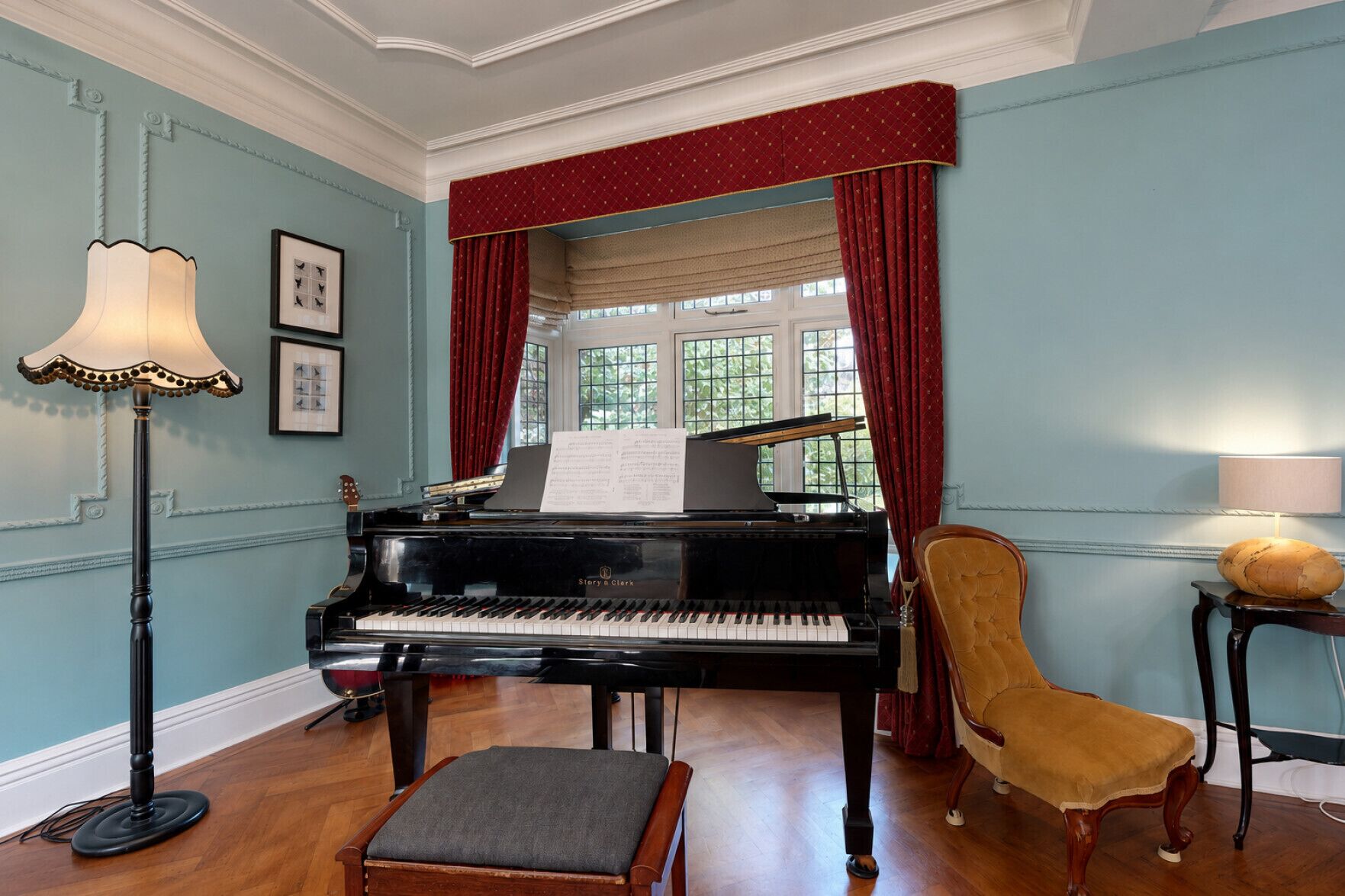 A grand piano in a room with a wooden floor, a chair, a lamp, and a window with curtains.