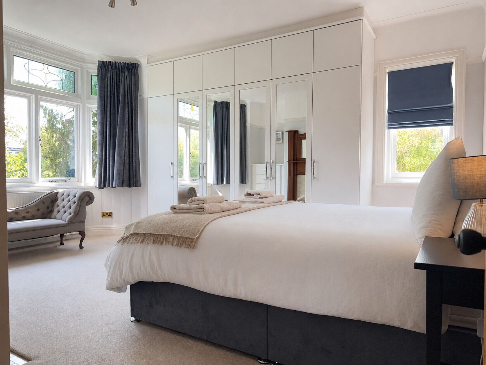 A bedroom with a large bed, a wardrobe, a sitting area with a chair, and a window with curtains.