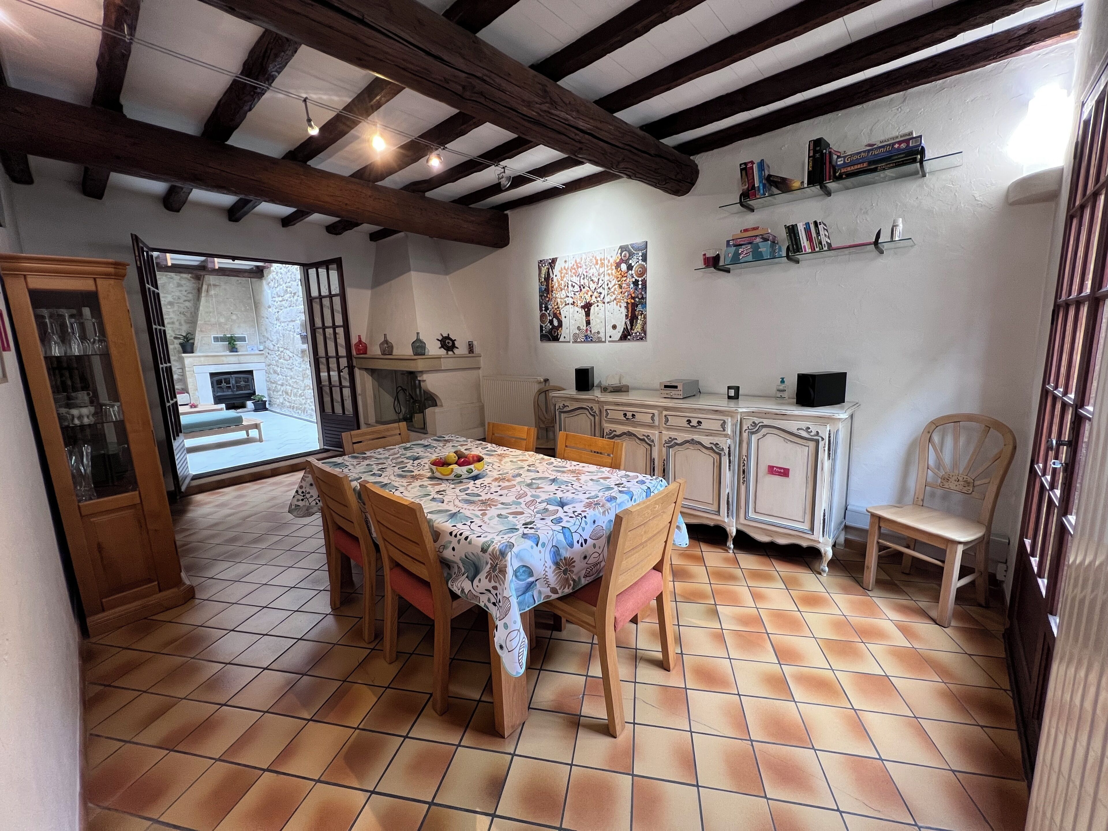 Un coin repas avec un sol carrelé, des chaises en bois, une table recouverte d’une nappe à motifs floraux et une cheminée.