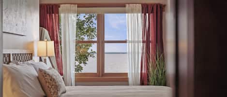 Une chambre avec une grande fenêtre, un lit avec des oreillers, une table de chevet avec une lampe, et une vue sur la mer.