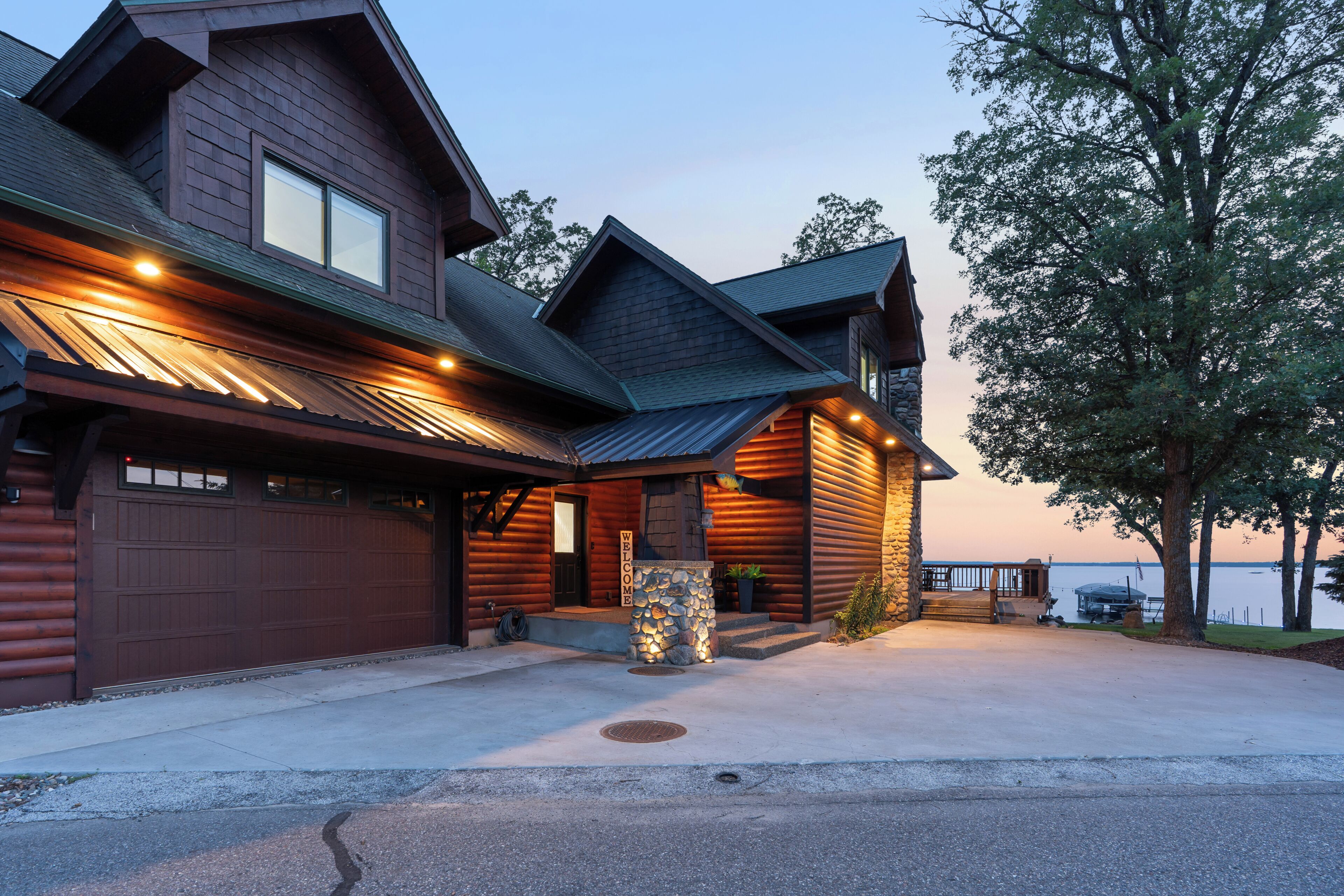 A large log cabin with a garage, a stone fireplace, and a deck overlooking a body of water.