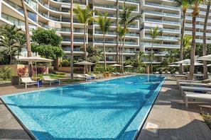 Ein Swimmingpool mit Liegestühlen und Sonnenschirmen, umgeben von Palmen und einem mehrstöckigen Gebäude.