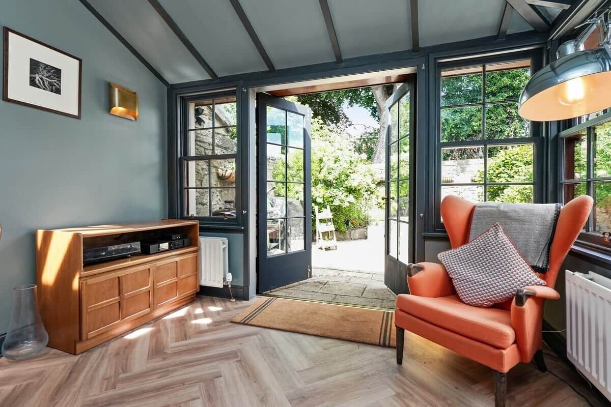 Una habitación con piso de espiga, un armario de madera, un sillón de cuero y un ventanal con vista a vegetación.