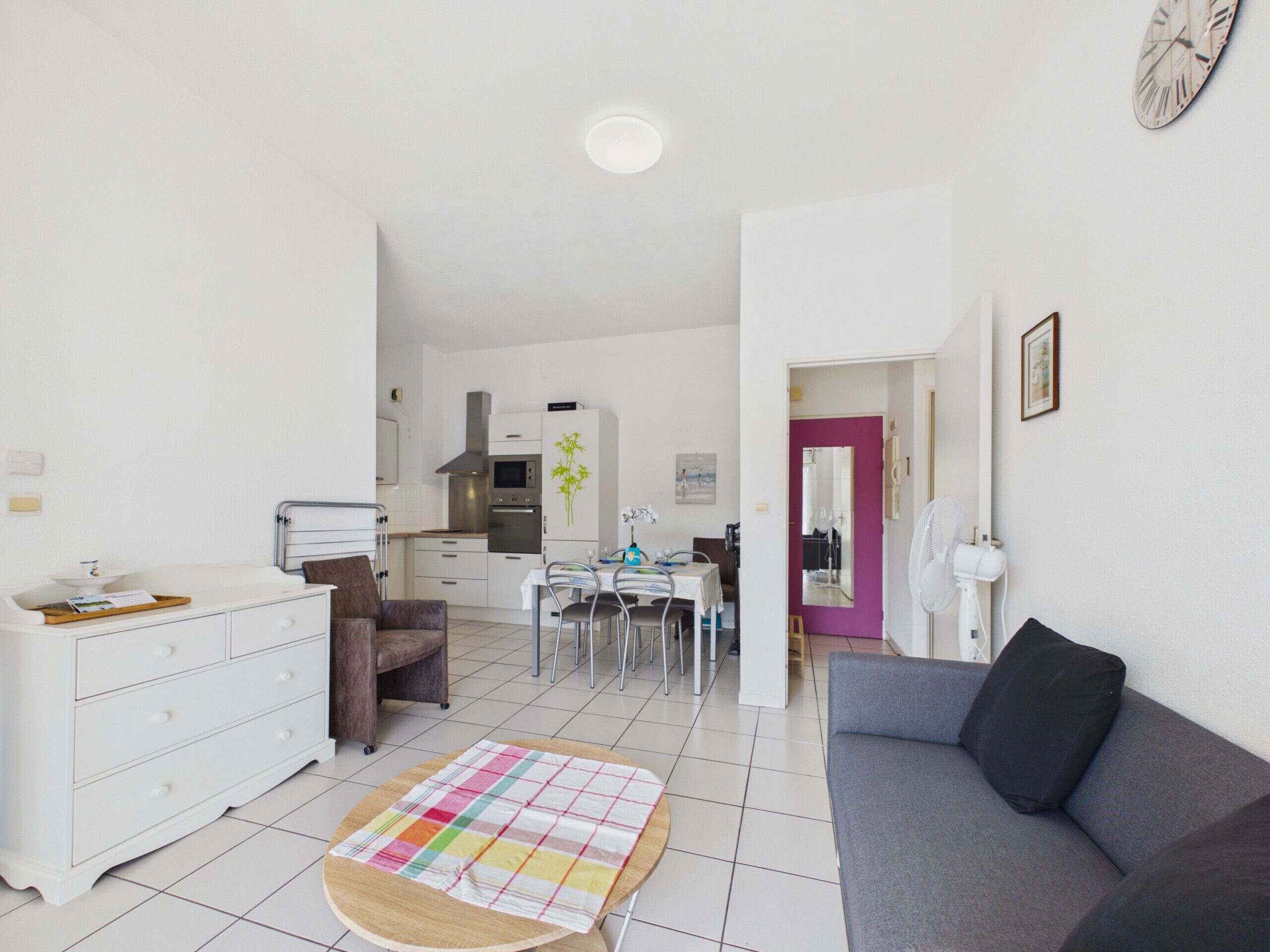 Un salon moderne avec un canapé gris, une table à manger avec des chaises, un coin cuisine avec des meubles blancs et un petit tapis rond.
