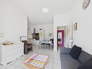 Ein modernes Wohnzimmer mit grauem Sofa, einem Esstisch mit Stühlen, einer Küchenzeile mit weißen Schränken und einem kleinen runden Teppich.