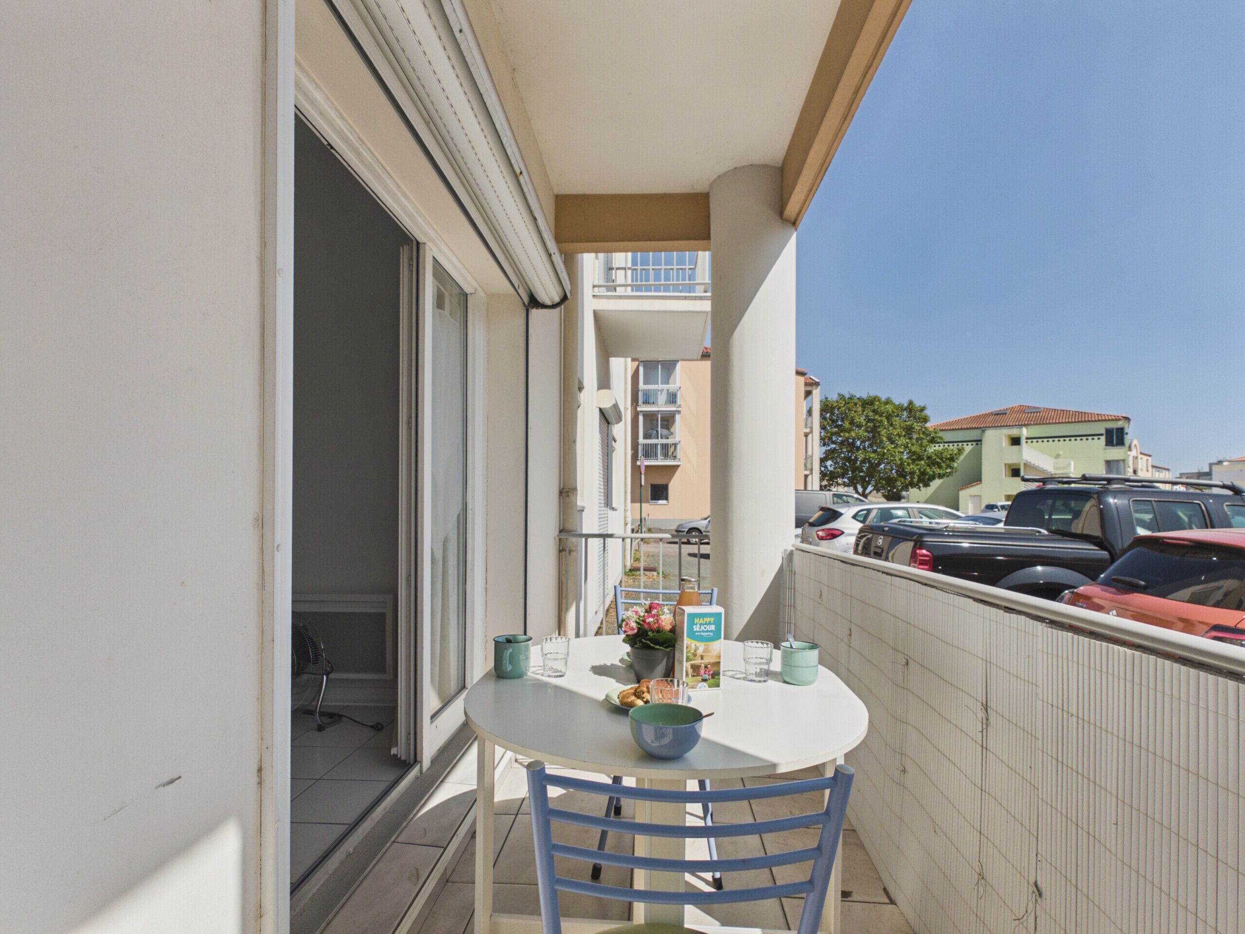 A balcony with a table and chairs, a potted plant, and a view of parked cars and buildings.