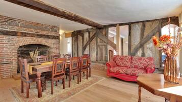 A room with a brick fireplace, wooden dining table with chairs, a red floral sofa, and a wooden coffee table.