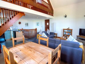 A spacious living area with a wooden staircase, a dining table with chairs, a sofa, a bookshelf, and a television. - Home Near Widemouth Bay (Bude)