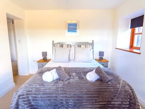 A bedroom with a large bed, two bedside tables, a nightstand, a window with a blind, and a painting on the wall. - Home Near Widemouth Bay (Bude)