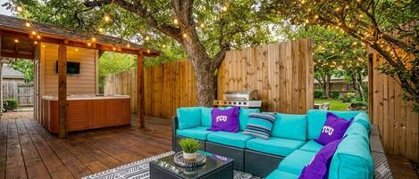 An outdoor patio with a wooden deck, a teal sofa with purple cushions, a barbecue grill, and string lights.