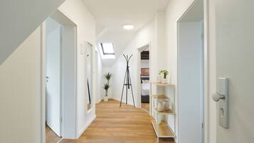 A hallway with wooden flooring, white walls, and a skylight. There are doors on both sides, a plant, and a shelf with decorative items.