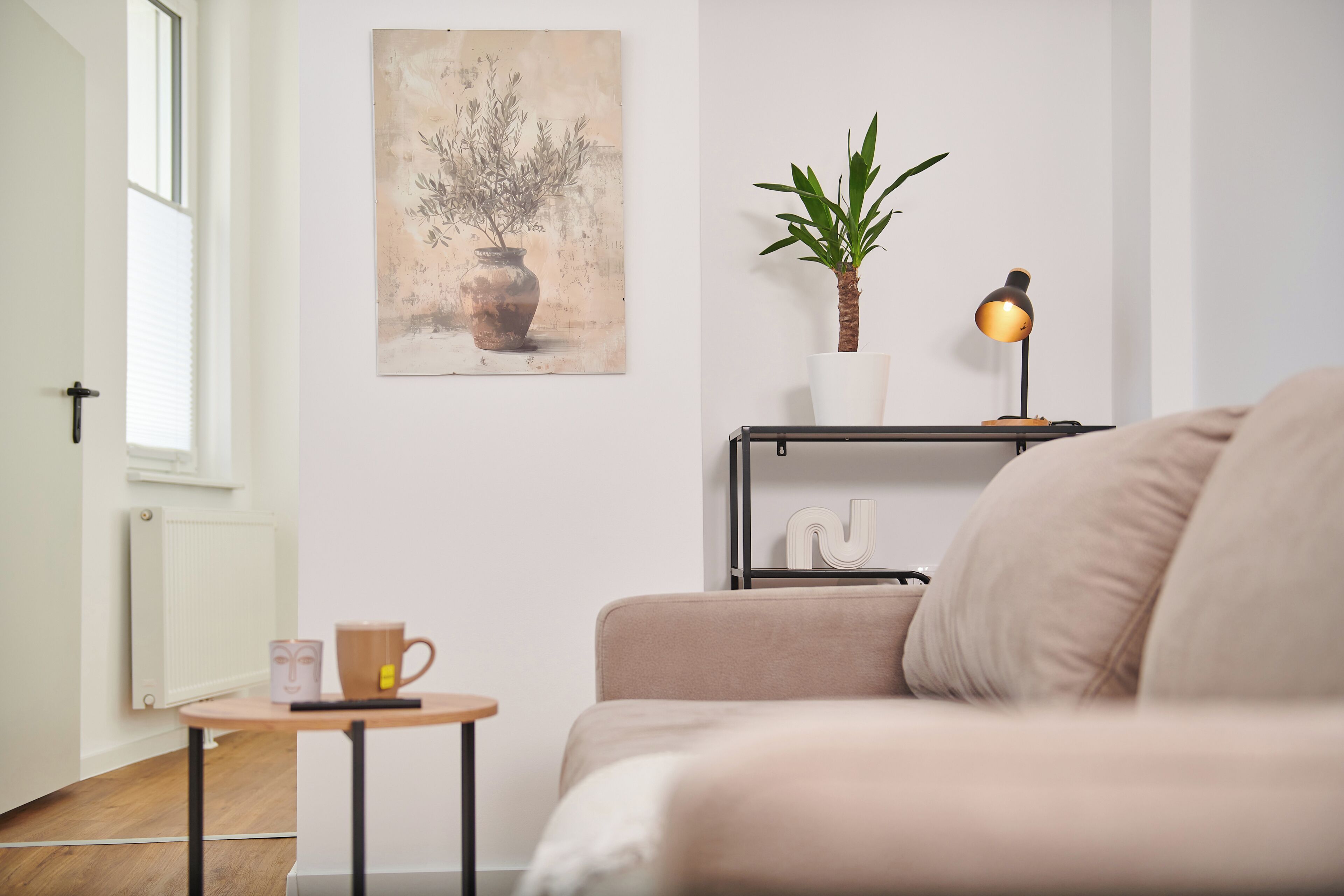 Ein minimalistisch eingerichtetes Wohnzimmer mit Sofa, Couchtisch, kleiner Pflanze, Lampe und einem Bild an der Wand.