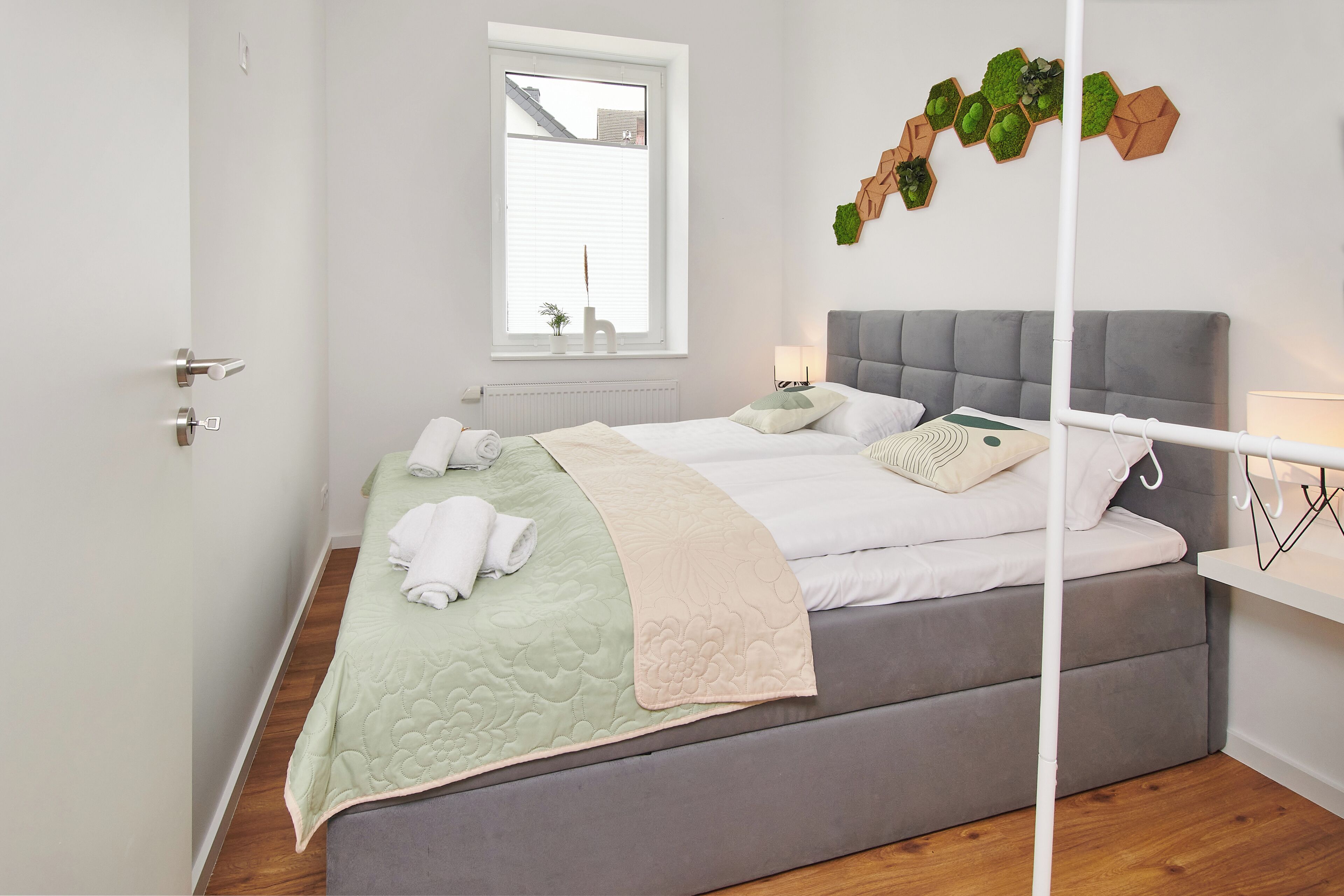Un dormitorio moderno con una cama tapizada en gris, una mesita de noche blanca y una ventana con persianas.