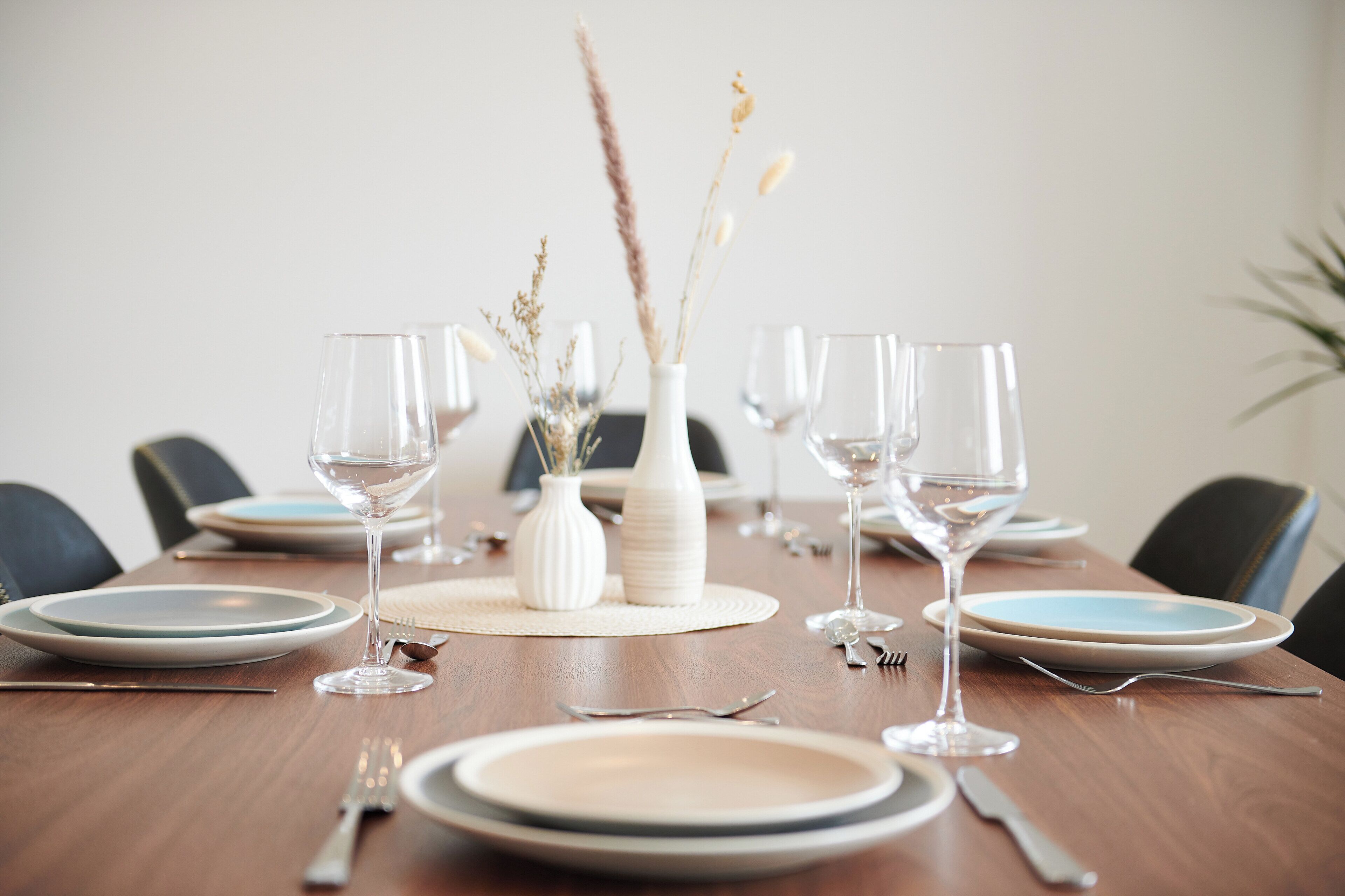 A dining table set with plates, glasses, and cutlery, with a vase of dried plants in the center.