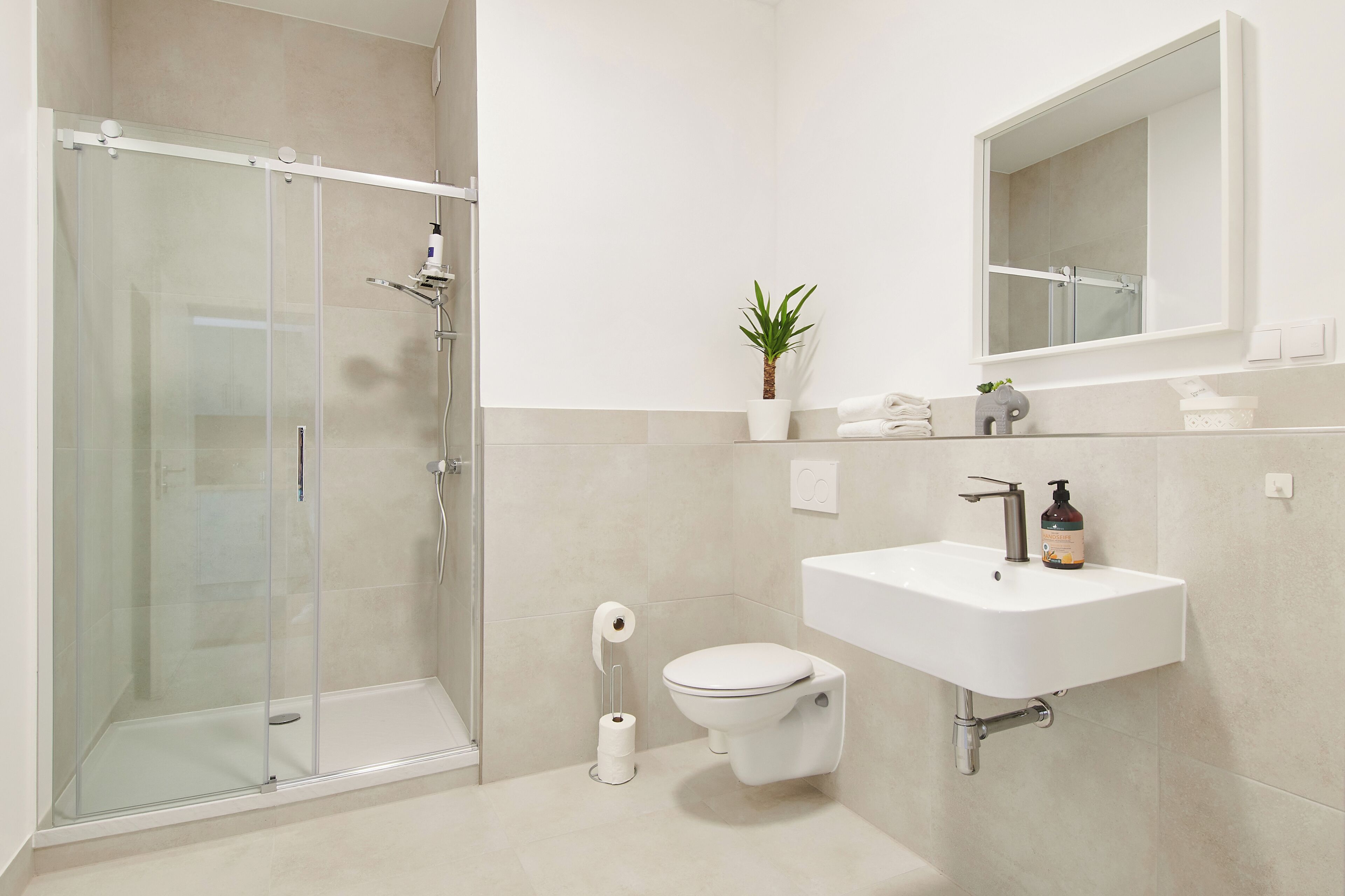 Ein modernes Badezimmer mit Duschkabine aus Glas, Toilette, Waschbecken mit Spiegel darüber und einer kleinen Topfpflanze.