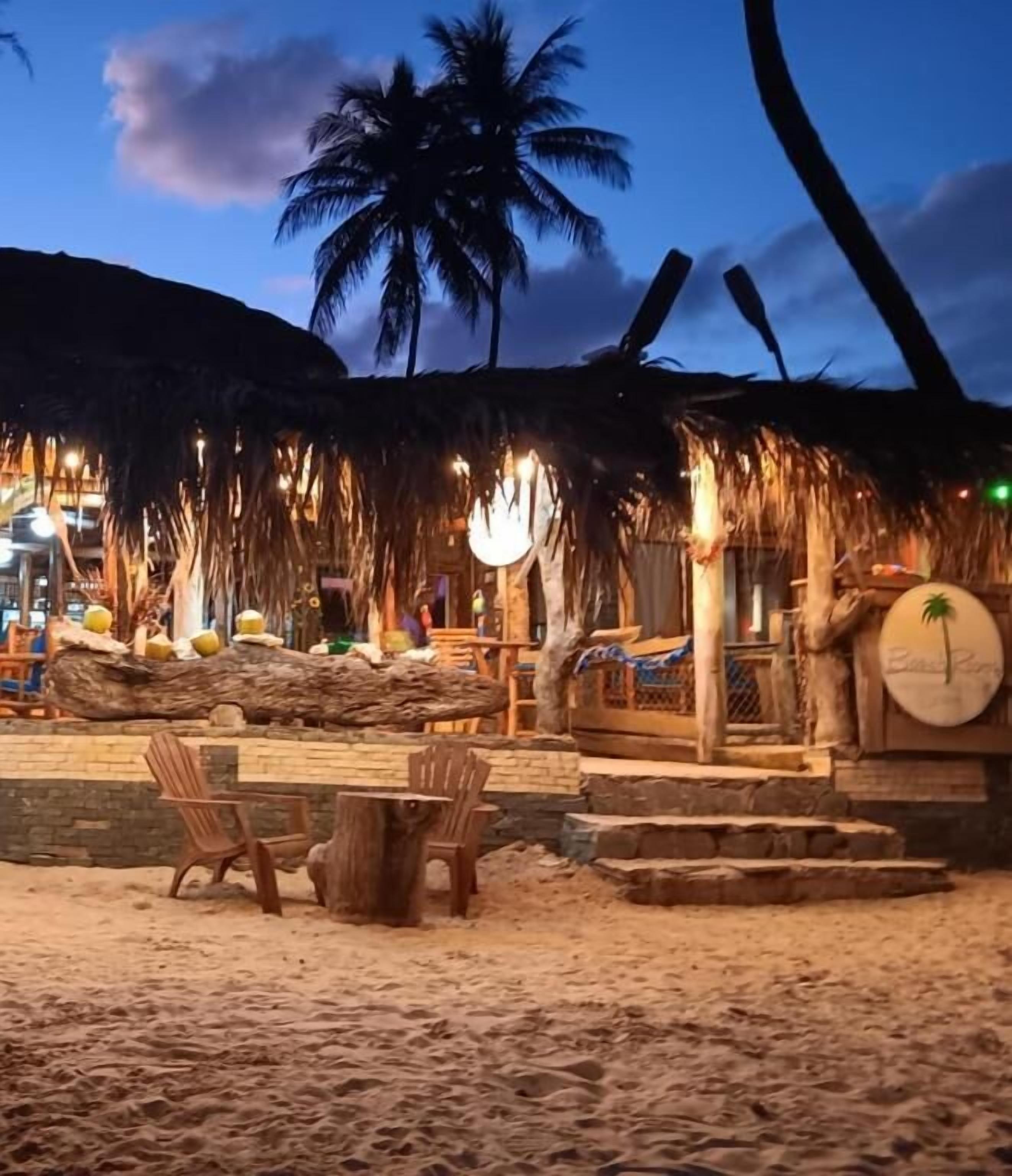 Een restaurant aan het strand met terras, palmbomen en een rieten dak.