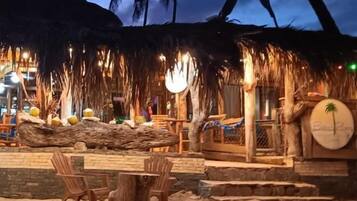 Een restaurant aan het strand met terras, palmbomen en een rieten dak.