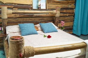 A rustic wooden bed with a woven headboard, two blue pillows, and a single red rose on a patterned bedspread.