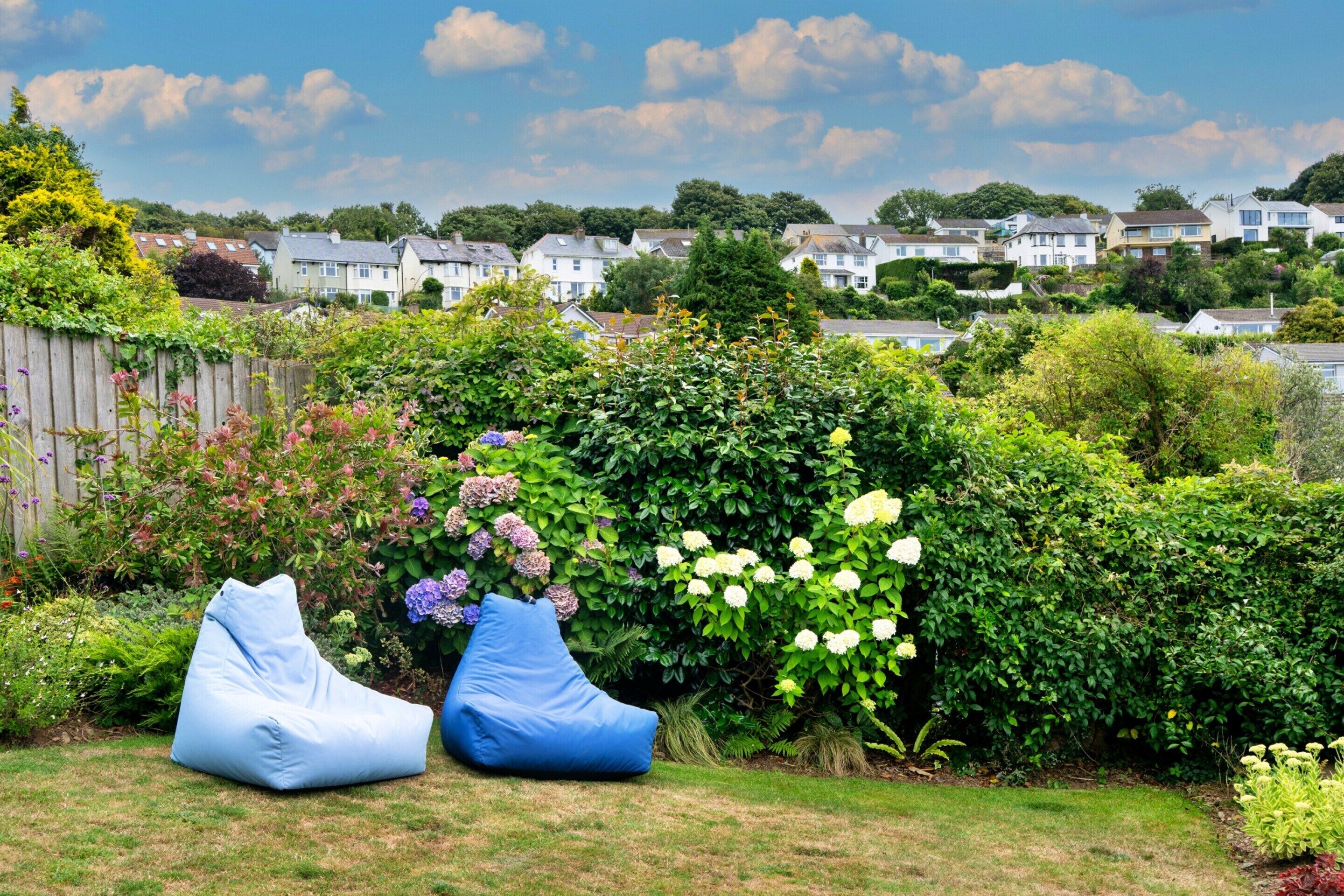 Un jardín con dos pufs azules, diversas plantas florecientes y una vista de casas al fondo.