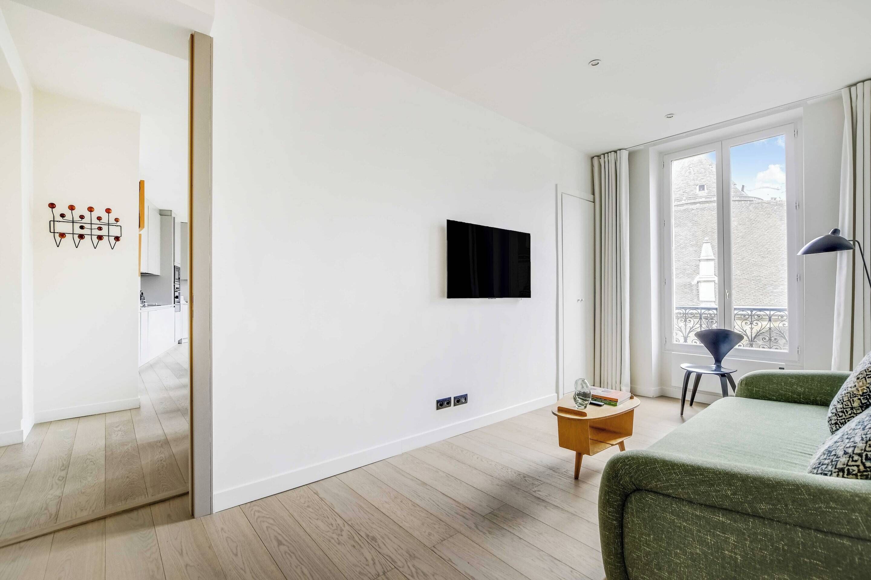 A modern living room with a flat-screen TV mounted on the wall, a green sofa, a small wooden side table, and a view of the outside through large windows.