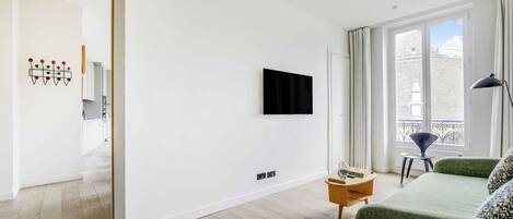 A modern living room with a flat-screen TV mounted on the wall, a green sofa, a small wooden side table, and a view of the outside through large windows.