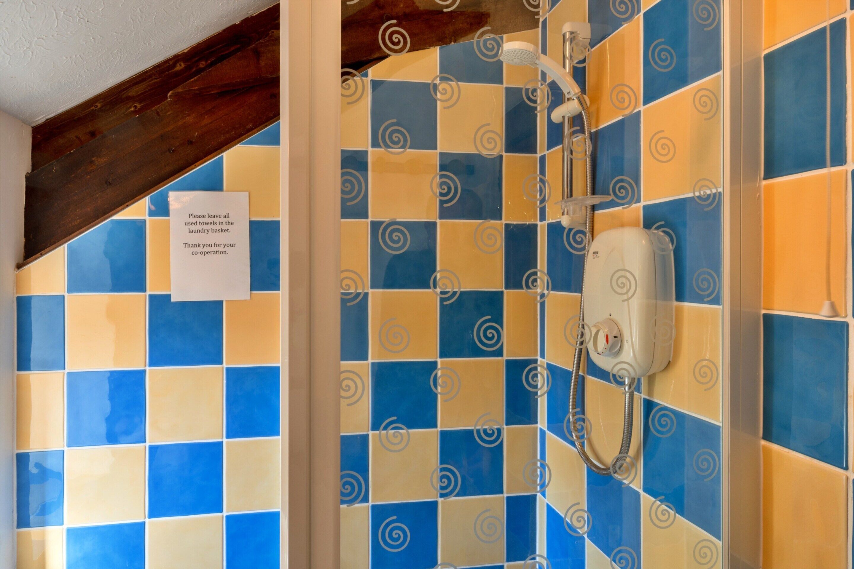 A bathroom with a checkered tile wall, a shower with a handheld showerhead, and a sign requesting towels to be left in the laundry basket.