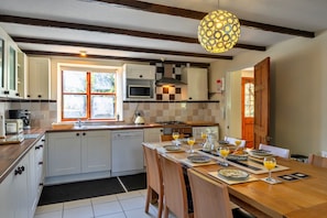 A kitchen with white cabinets, a microwave, a kettle, and a dining table set with plates and glasses. - Spacious Devon Farmhouse (Exeter)