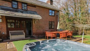 A hot tub in a backyard with a brick house, a wooden deck, and a patio table with chairs. - Spacious Devon Farmhouse (Exeter)