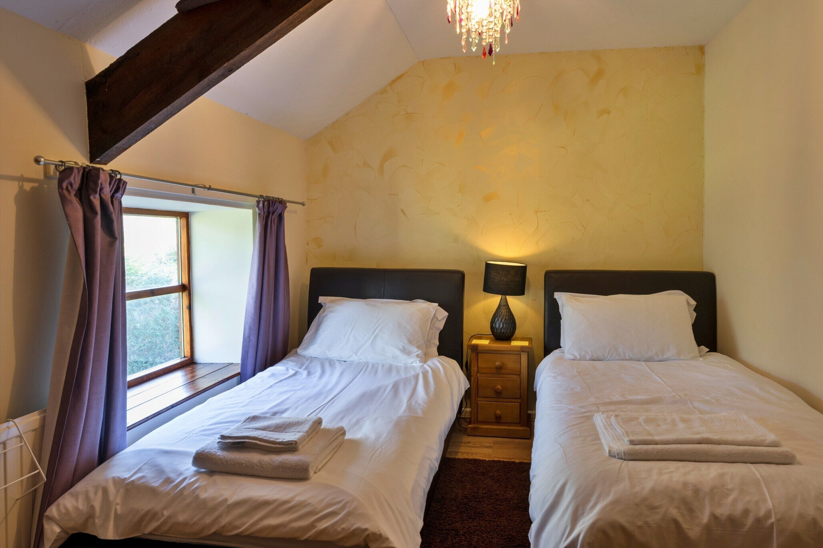 A hotel room with two beds, a window with curtains, a chandelier, and a nightstand with a lamp.