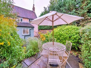 A garden patio with a table and chairs, surrounded by lush greenery and a house in the background. - Charming Country Cottage Near Bath (Trowbridge)