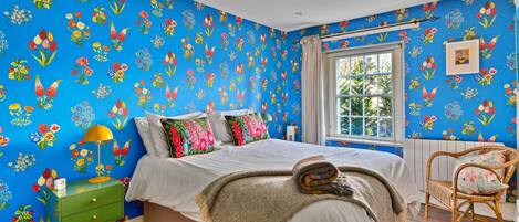 A bedroom with a floral wallpaper, a bed with white bedding, a green nightstand, a wicker chair, and a window with a view.