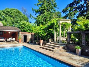 Ein Swimmingpool mit einer Steinterrasse, einer überdachten Lounge-Ecke und einer Treppe zu einer Pergola.