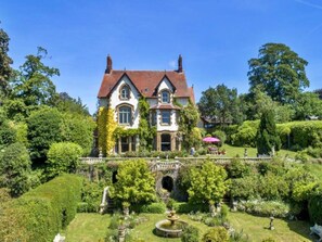 Ein großes, zweistöckiges Haus mit einer symmetrischen Fassade, einem zentralen Eingang und einem gepflegten Garten mit einem Brunnen.