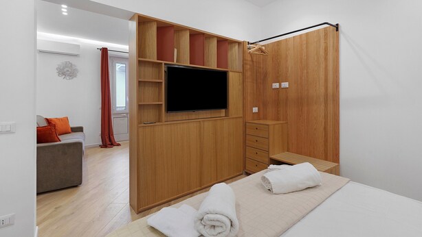 Une chambre moderne avec une armoire en bois, un téléviseur à écran plat, un lit avec du linge de maison blanc et un petit coin salon comprenant un canapé et des coussins rouges.