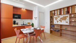 Une cuisine moderne avec des meubles de couleur orange, un coin repas avec des chaises assorties et une bibliothèque décorée sur le thème du voyage.