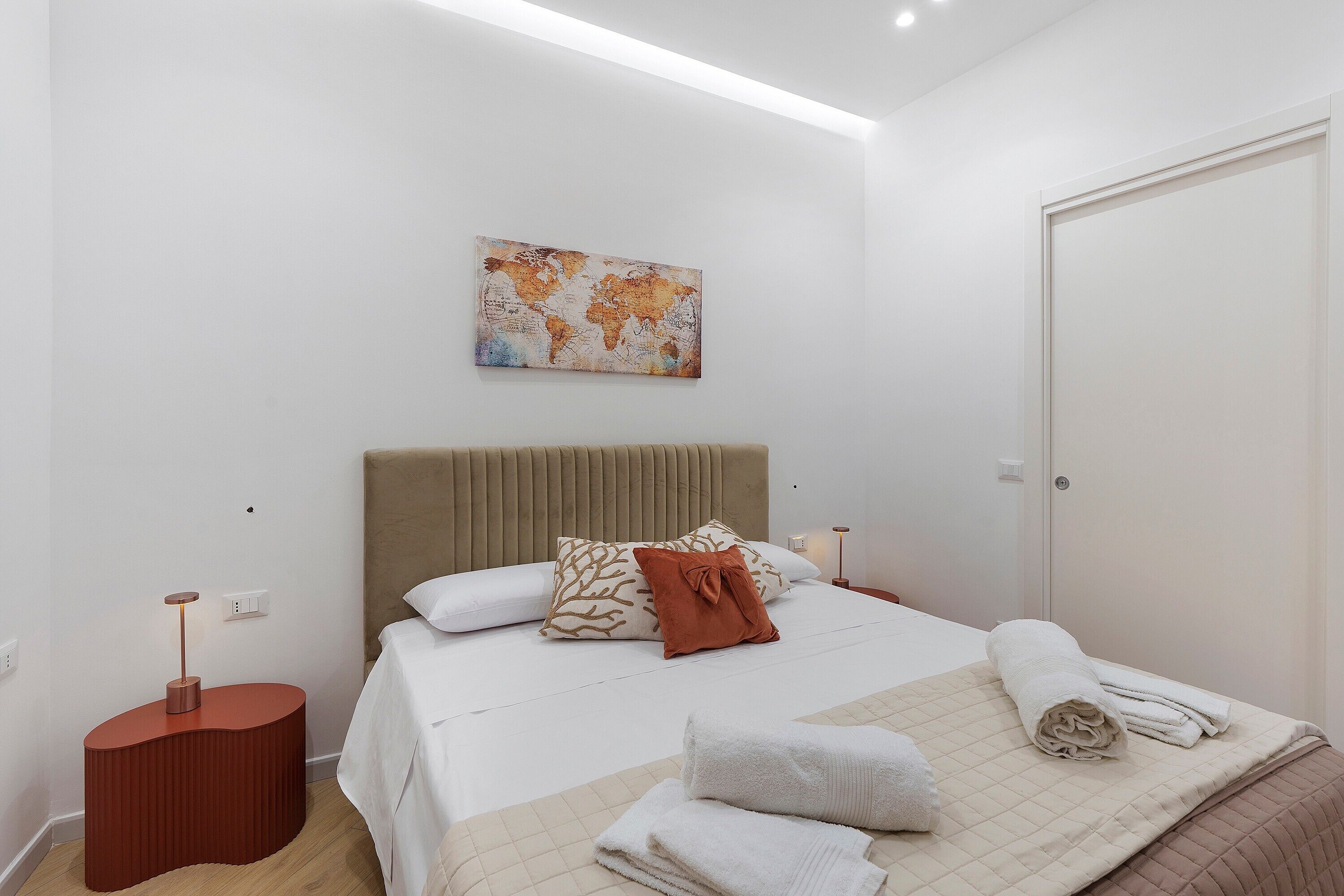 A modern bedroom with a bed, pillows, a bedside table, a floor lamp, and a world map artwork on the wall.