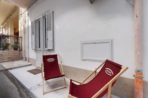 Zwei Holzstühle mit roten Kissen vor einem Gebäude mit weißer Wand und einer Tür mit Fensterläden.